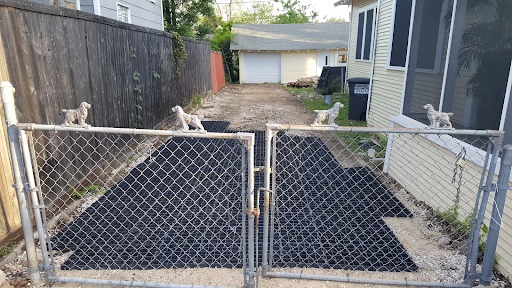 An image of a yellow house and wire fence bordered by a dirt driveway partially covered with black Ecoraster tiles. The image was taken mid-installation of permeable pavement.