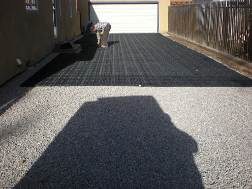 A gravel driveway being paved with Ecoraster. Half of the driveway is grey gravel, while on the second half a man is bent over, installing black Ecoraster tiles. A fence runs along the right-hand edge of the driveway.