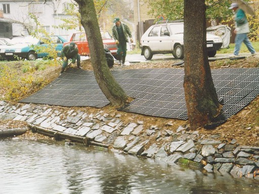 A riverbank seen from across a small river. A foundation of permeable pavement has been laid, and people mill around adjusting it. 