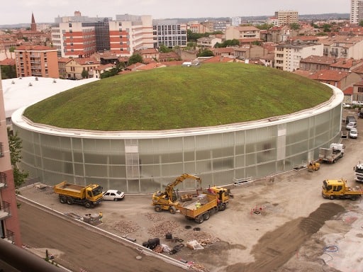 An aerial view of an oval building in a city. The terra-cotta colours of the city in the background contrast with the oval building in the foreground, which has a rounded green roof covered in plant life. 